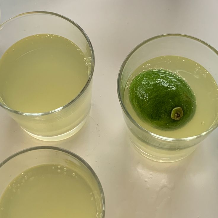 three glasses filled with green liquid next to an avocado on a white surface