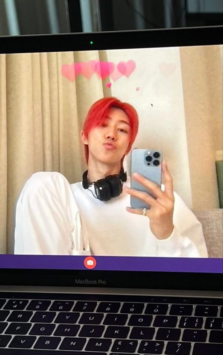 a person with red hair and headphones holding up a cell phone in front of a laptop