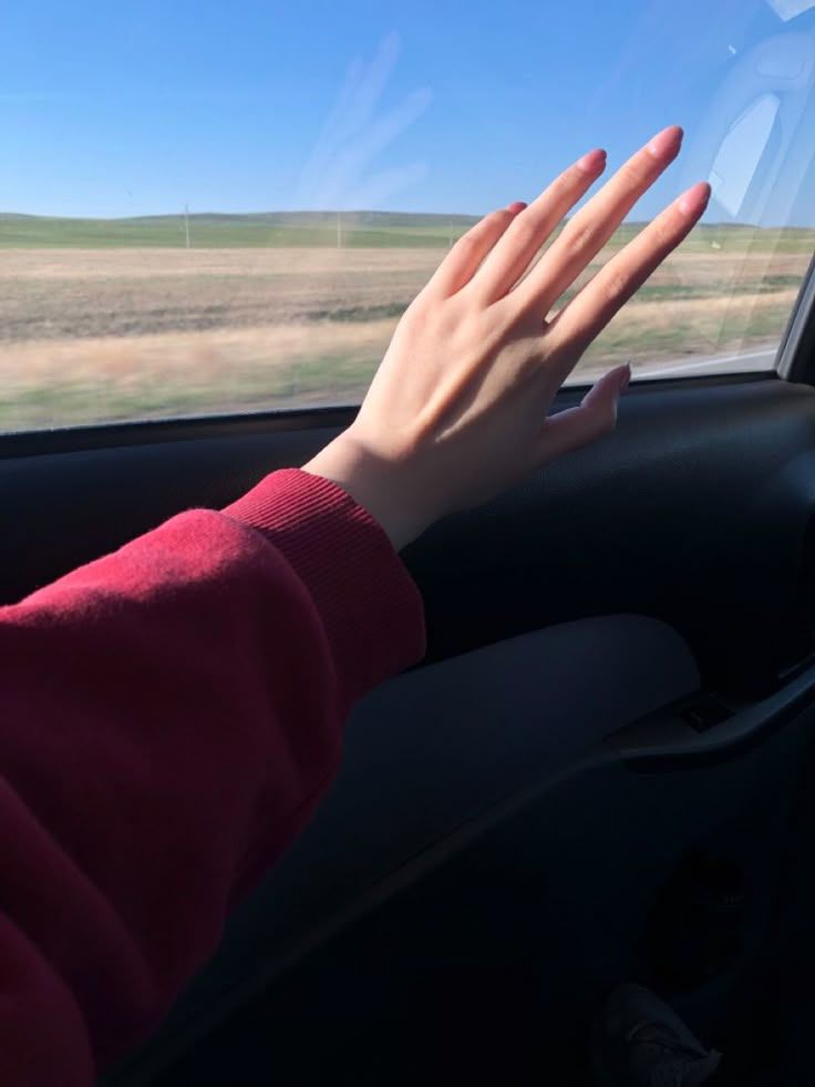 a person holding their hand out the window of a car on a road with grass and blue sky in the background