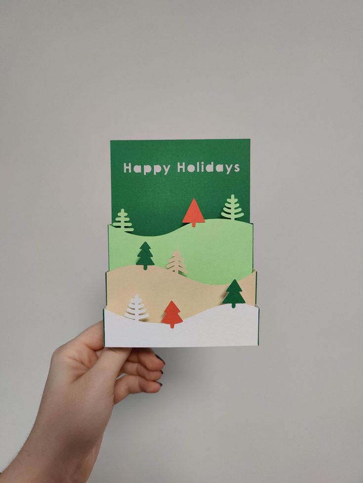 someone holding up a card that says happy holidays with trees and mountains in the background