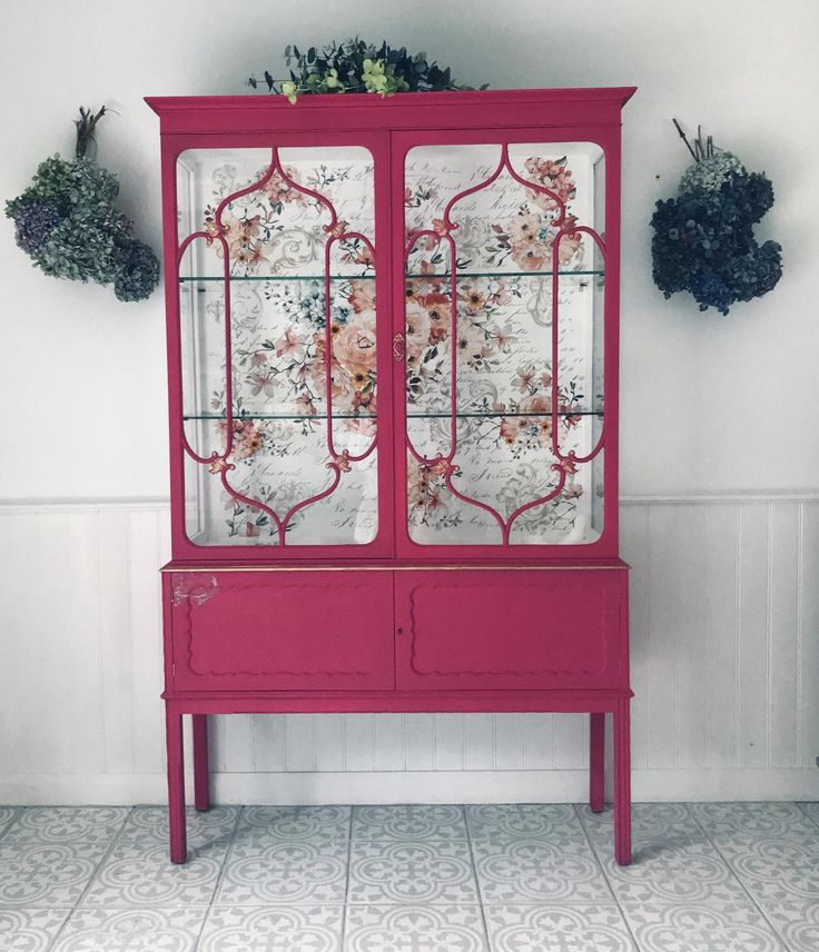 a pink china cabinet with glass doors and flowers on the top, against a white wall