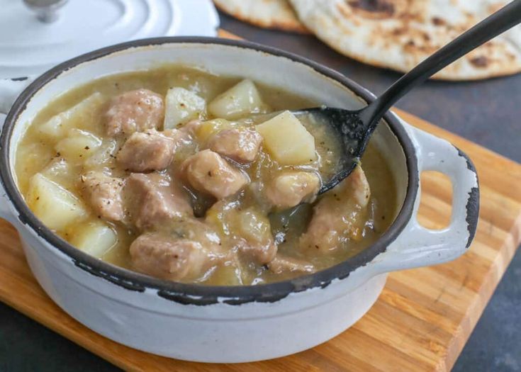 a white pot filled with meat and potatoes on top of a wooden cutting board next to pita bread