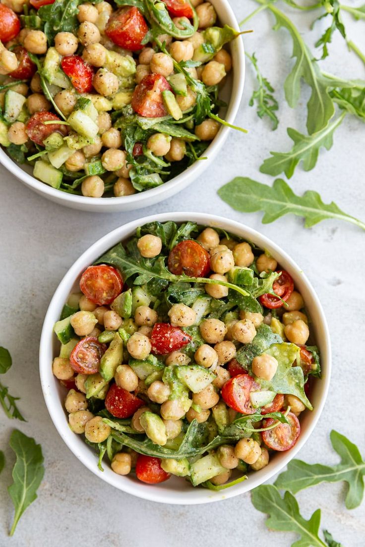 two bowls filled with chickpeas, tomatoes and lettuce on top of a white table