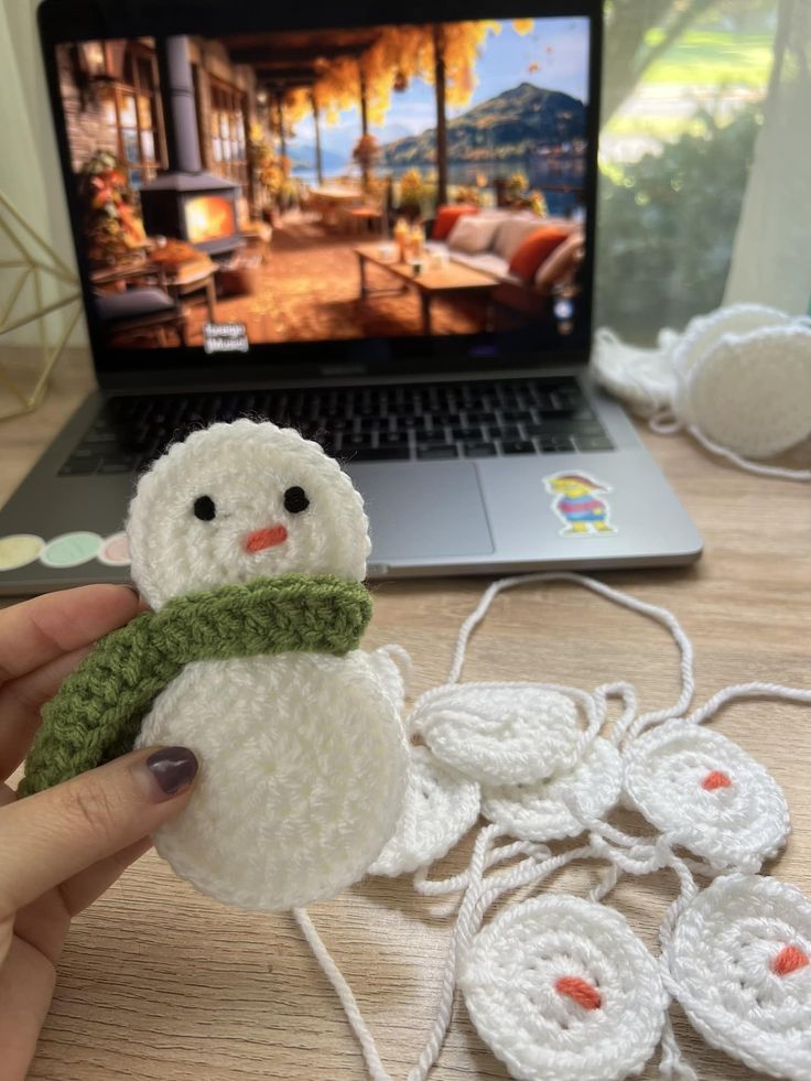 someone is holding a small snowman in front of a laptop on a table with other crocheted items