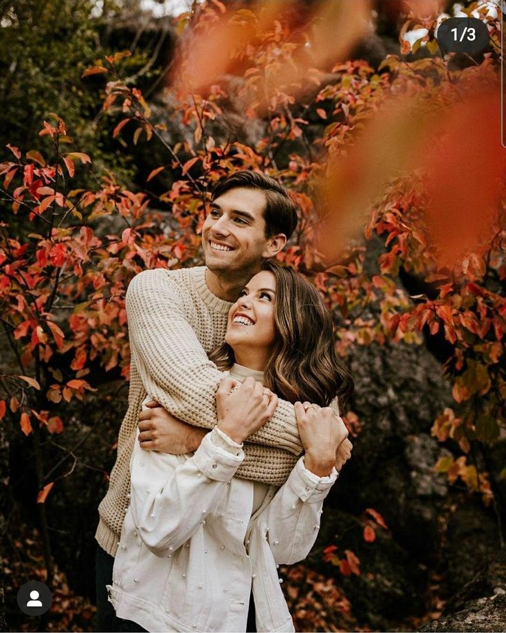 a man and woman hugging each other in front of trees with red leaves on them