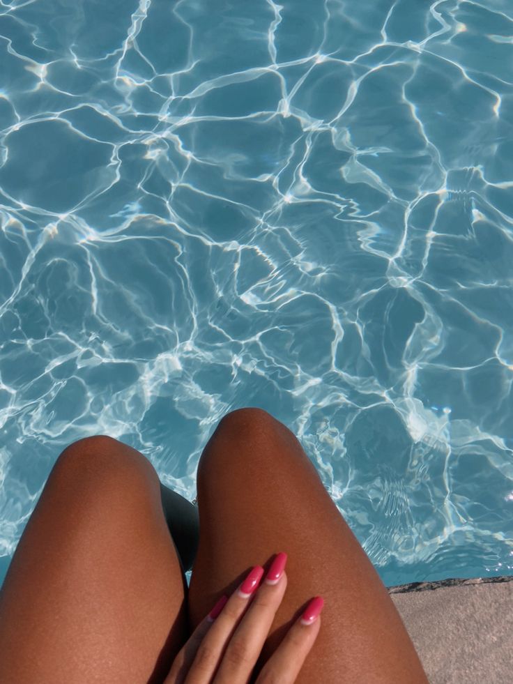 a woman's legs with pink nail polish sitting in front of a swimming pool