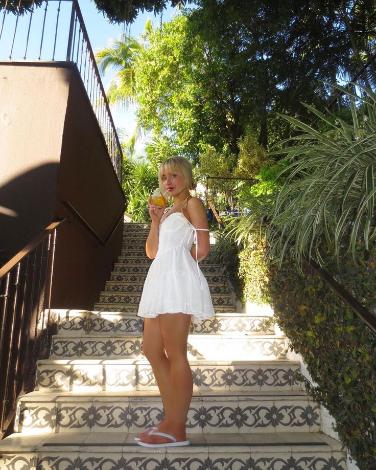 a woman in white dress standing on steps eating an orange and looking at the camera