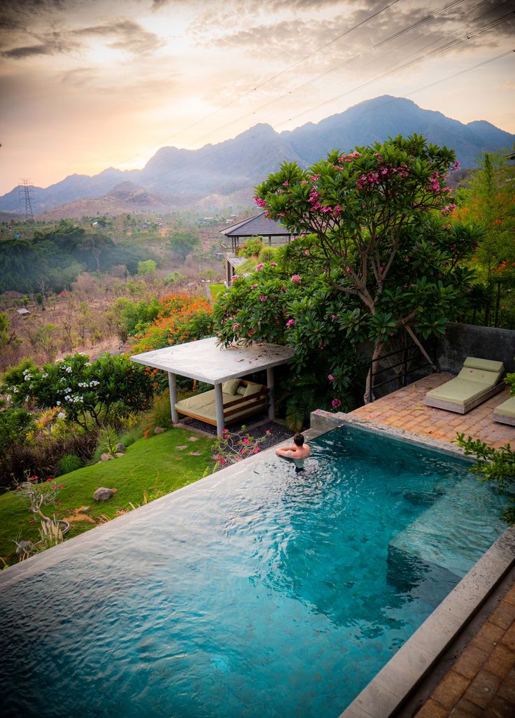 an outdoor swimming pool surrounded by lush greenery and mountains in the distance, with a person jumping into it