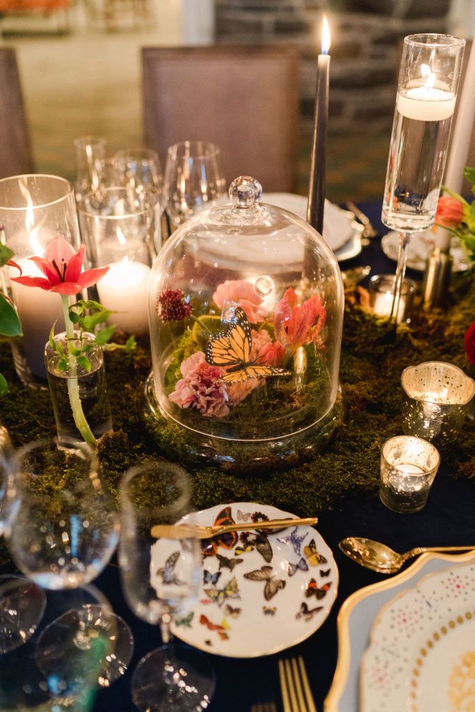 the table is set with candles, plates and flowers in a glass dome on top of moss