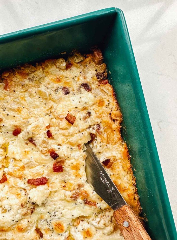 a green casserole dish filled with cheese and bacon, topped with a wooden spatula