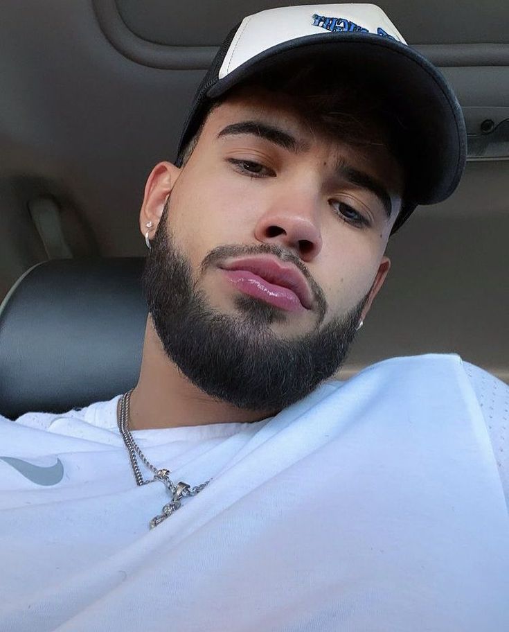 a man with a beard wearing a white shirt and black hat sitting in the back seat of a car