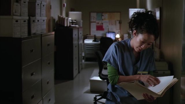 a woman in scrubs is looking through a folder while sitting at a desk with filing cabinets behind her