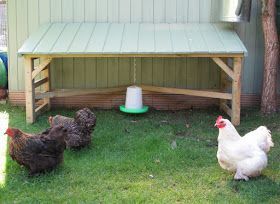 three chickens are standing in the grass near a wooden structure and a green bird feeder