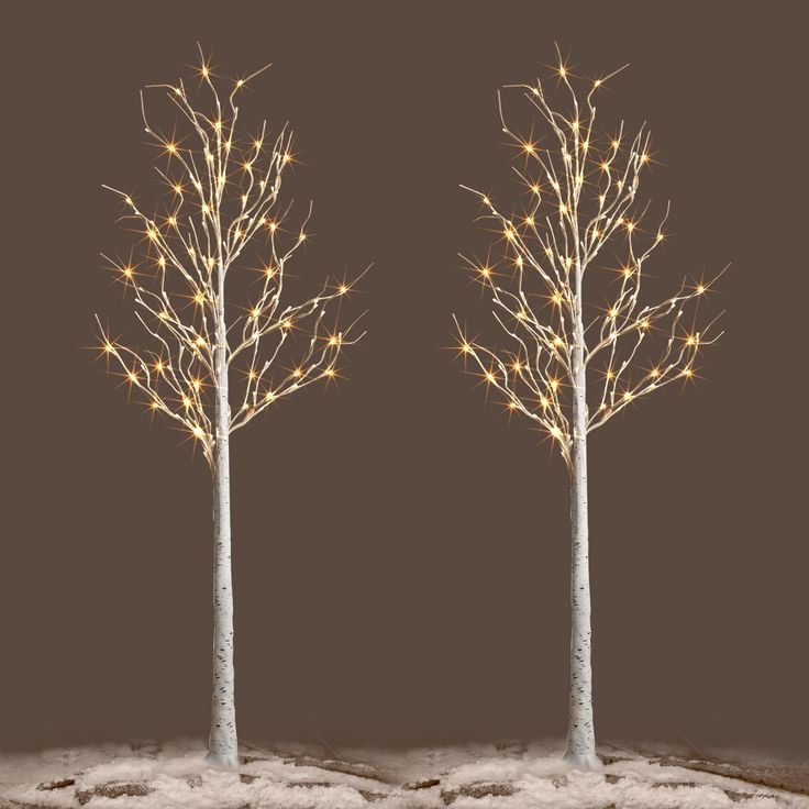 two white trees with lights on them in the middle of snow covered ground, against a dark background