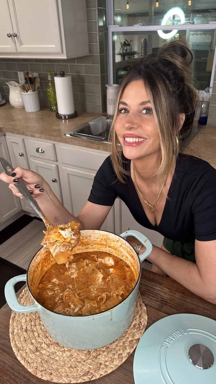 a woman sitting at a table in front of a large pot filled with food and holding a spoon