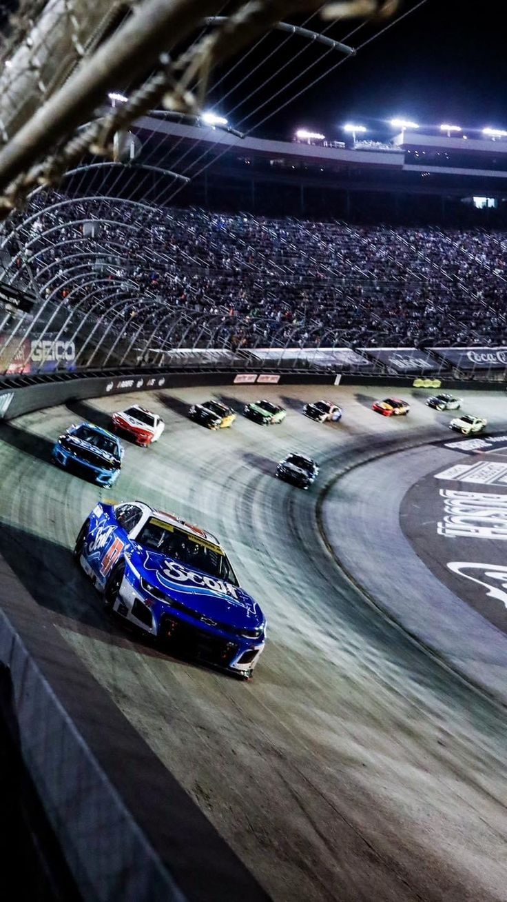 cars racing around a track in front of an audience at a nascar race event, with the lights on