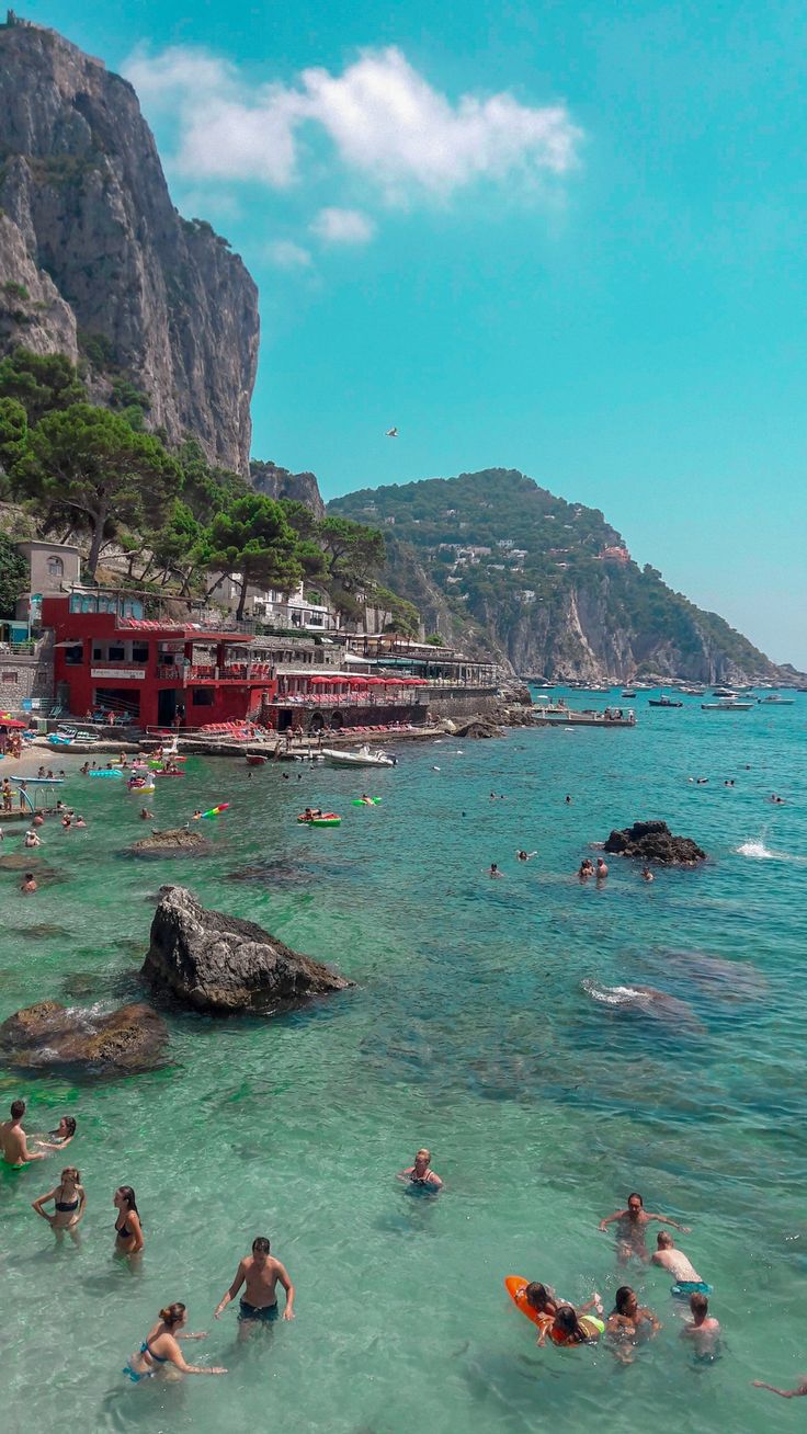 many people are swimming in the clear blue water near some cliffs and houses on the shore