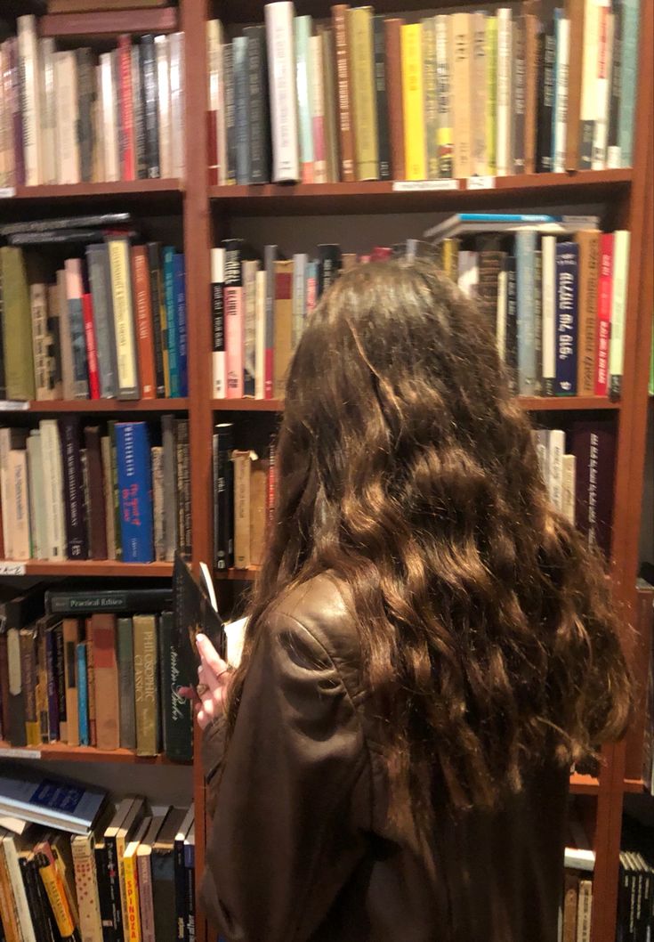 a woman standing in front of a bookshelf holding a cell phone and looking at it