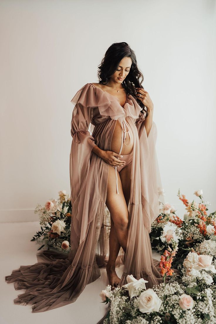 a pregnant woman standing in front of flowers wearing a flowing dress and holding her belly