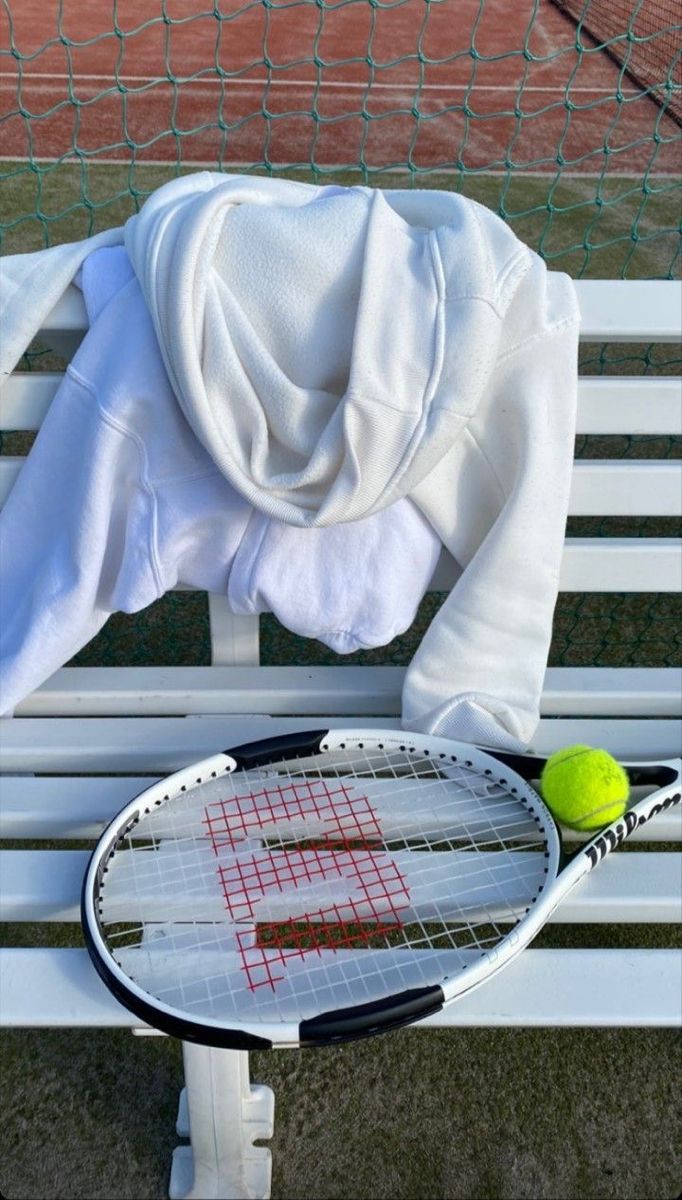 a tennis racket and ball sitting on a bench next to a net with a hoodie over it