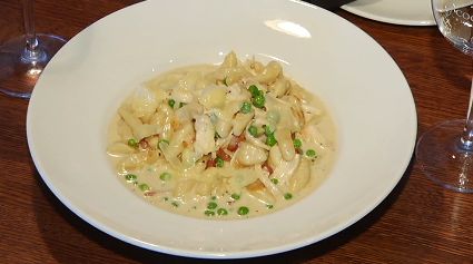 a white bowl filled with pasta and peas on top of a wooden table next to wine glasses