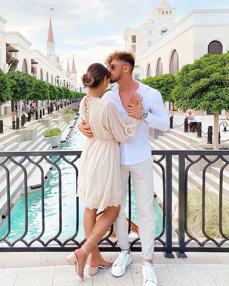 a man and woman standing next to each other in front of a fountain with buildings behind them