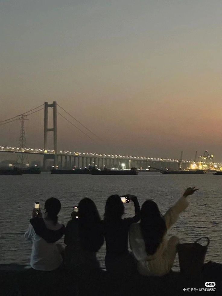 four people are sitting on a wall looking at the water with a bridge in the background