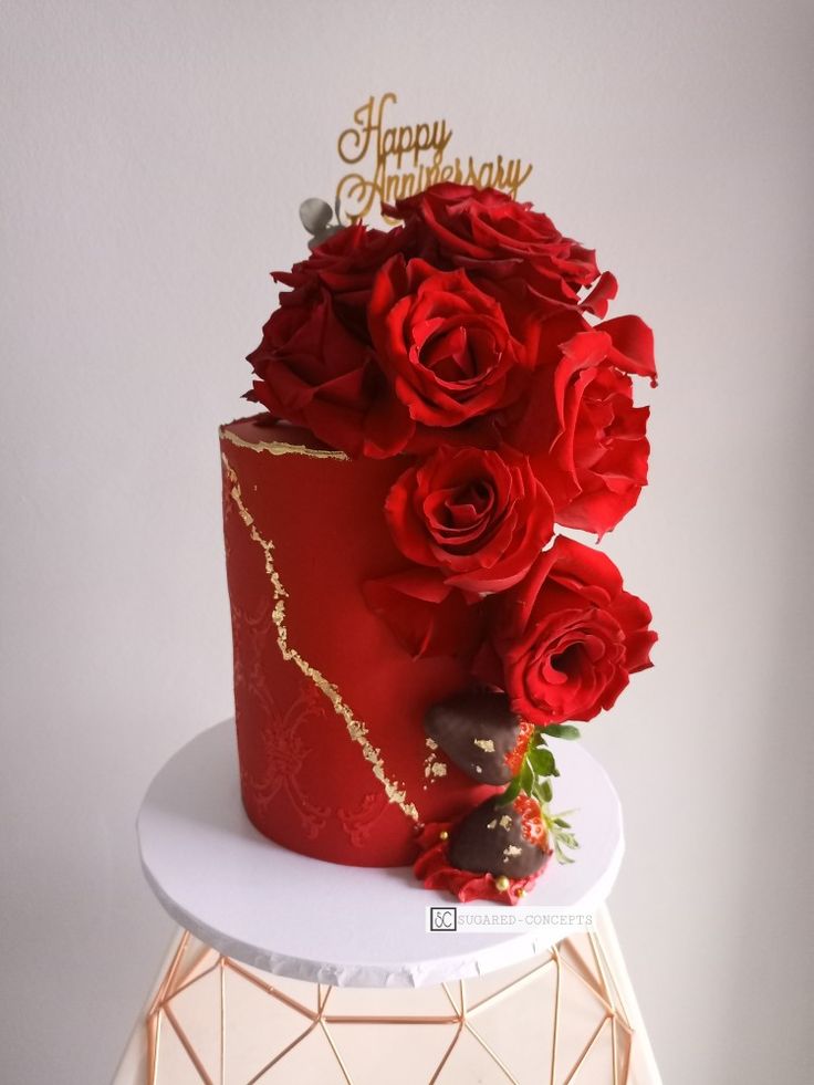 a red and gold birthday cake with roses on the top is sitting on a white table