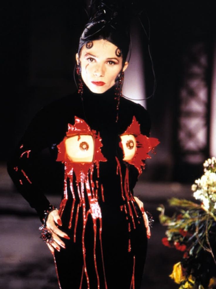 a woman in a black dress with red and orange decorations on her body, standing next to flowers