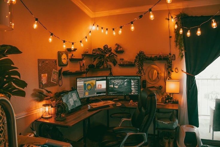 a computer desk with two monitors on it and lights strung from the ceiling in an office