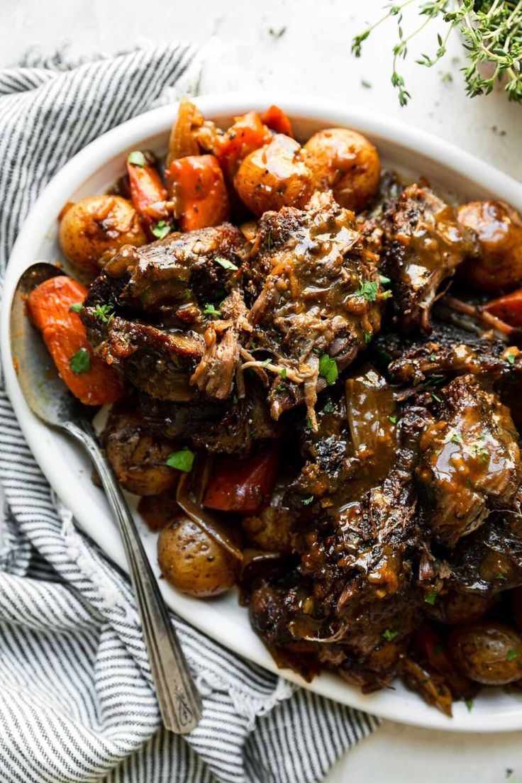 a white bowl filled with meat and potatoes on top of a blue striped towel next to a fork