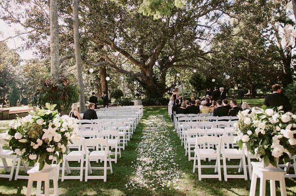 an outdoor ceremony setup with white chairs and flowers on the aisle, surrounded by trees