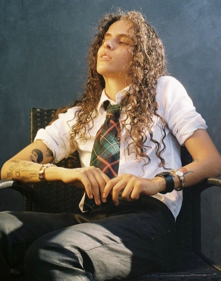 a woman with long curly hair sitting in a chair wearing a tie and looking off to the side