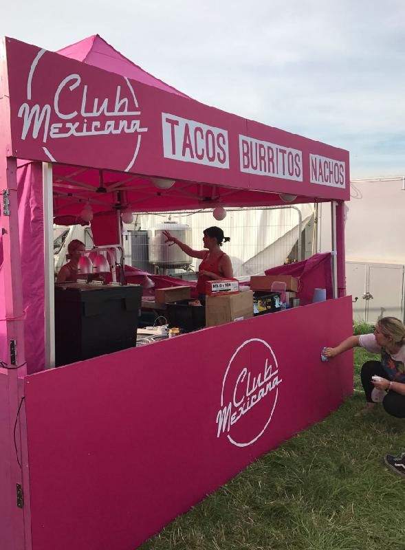 a woman sitting on the ground in front of a pink food cart with tacos written on it