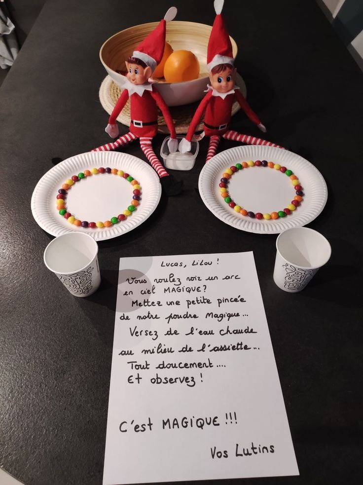 two elfs sitting at a table with paper plates and candy on it, next to a note