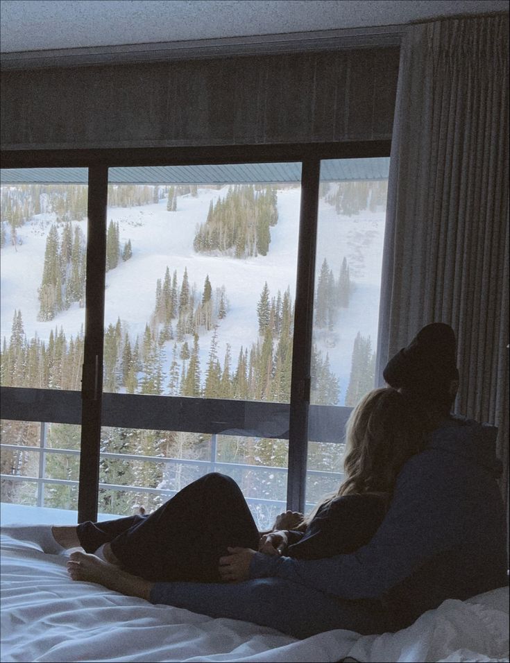 two people sitting on a bed looking out the window at snow covered mountains and trees