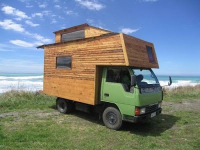 a green truck parked on top of a grass covered field next to the ocean with a wooden cabin attached to it's roof