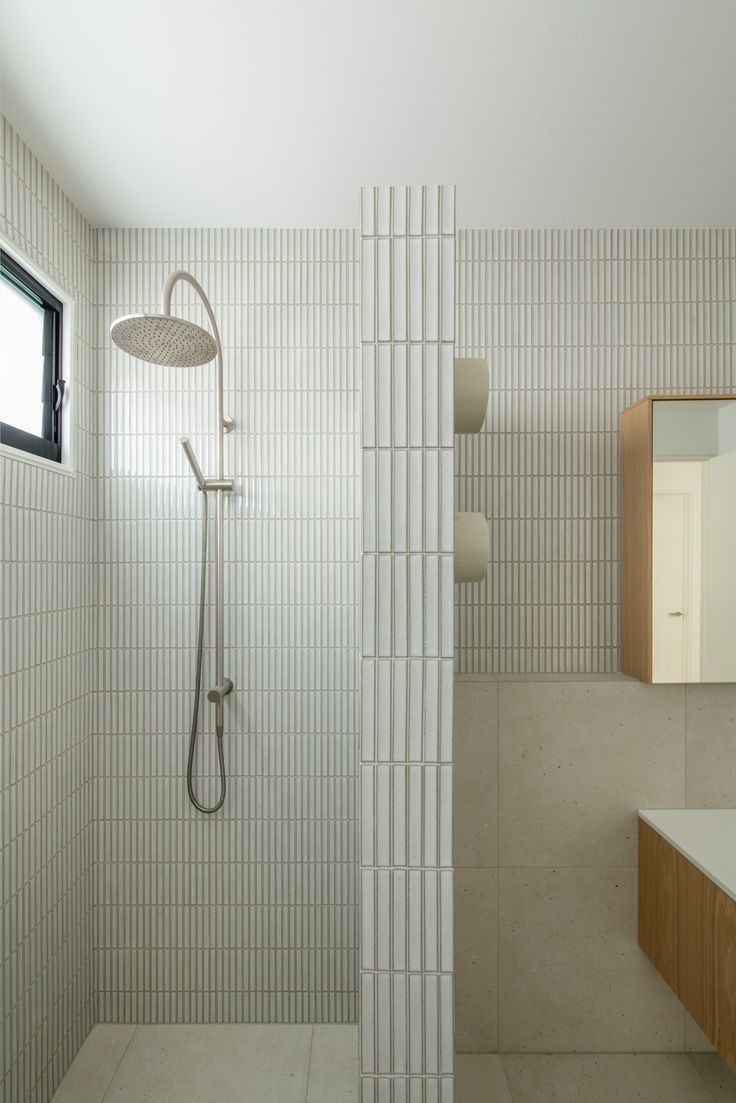 a white tiled bathroom with a shower head and mirror above the sink, in front of a window