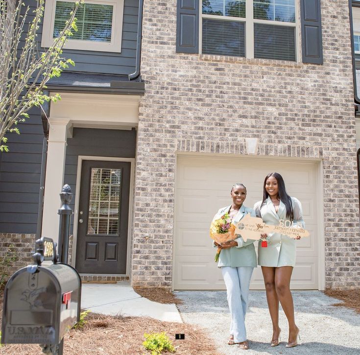 two women standing in front of a brick house holding a sign that says welcome home