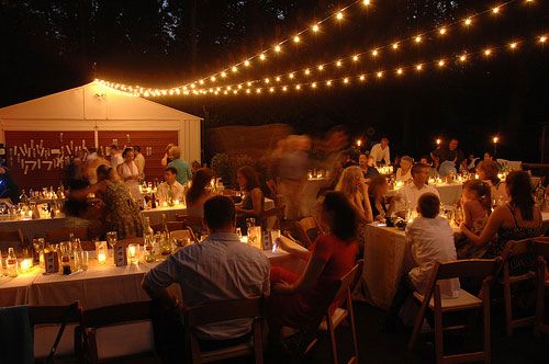 a group of people sitting at tables with candles in front of them and lights strung from the ceiling