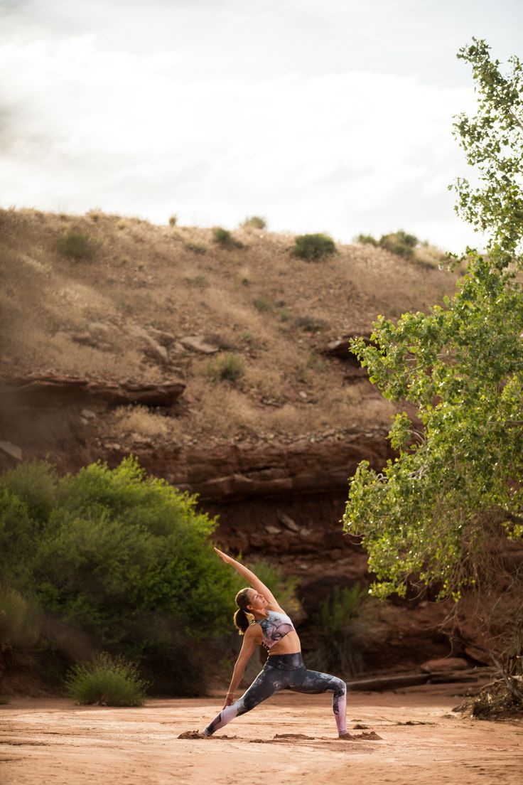 a woman is doing yoga outside in the dirt near some trees and bushes on a hill