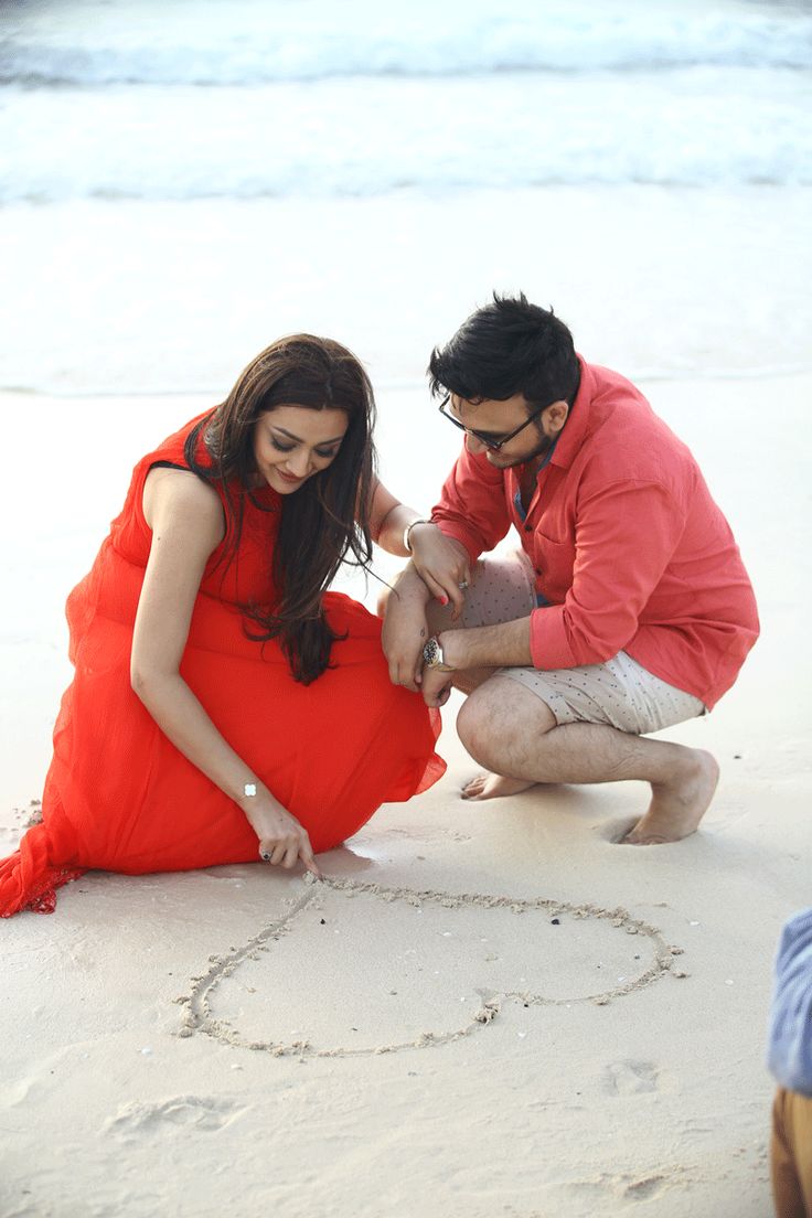 a man kneeling down next to a woman in a red dress on the beach with a heart drawn in the sand