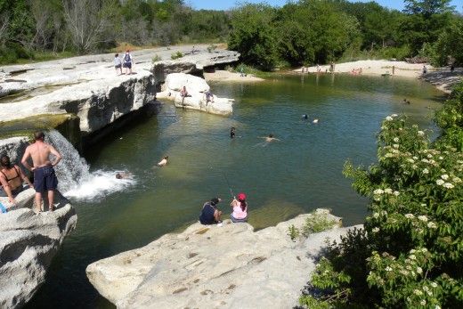 several people are swimming in the water near some rocks and boulders, while others stand around them