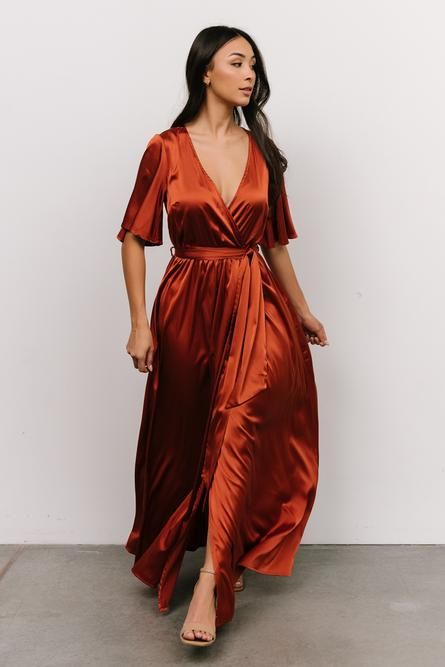 a woman wearing an orange satin wrap dress and matching heels, standing in front of a white wall