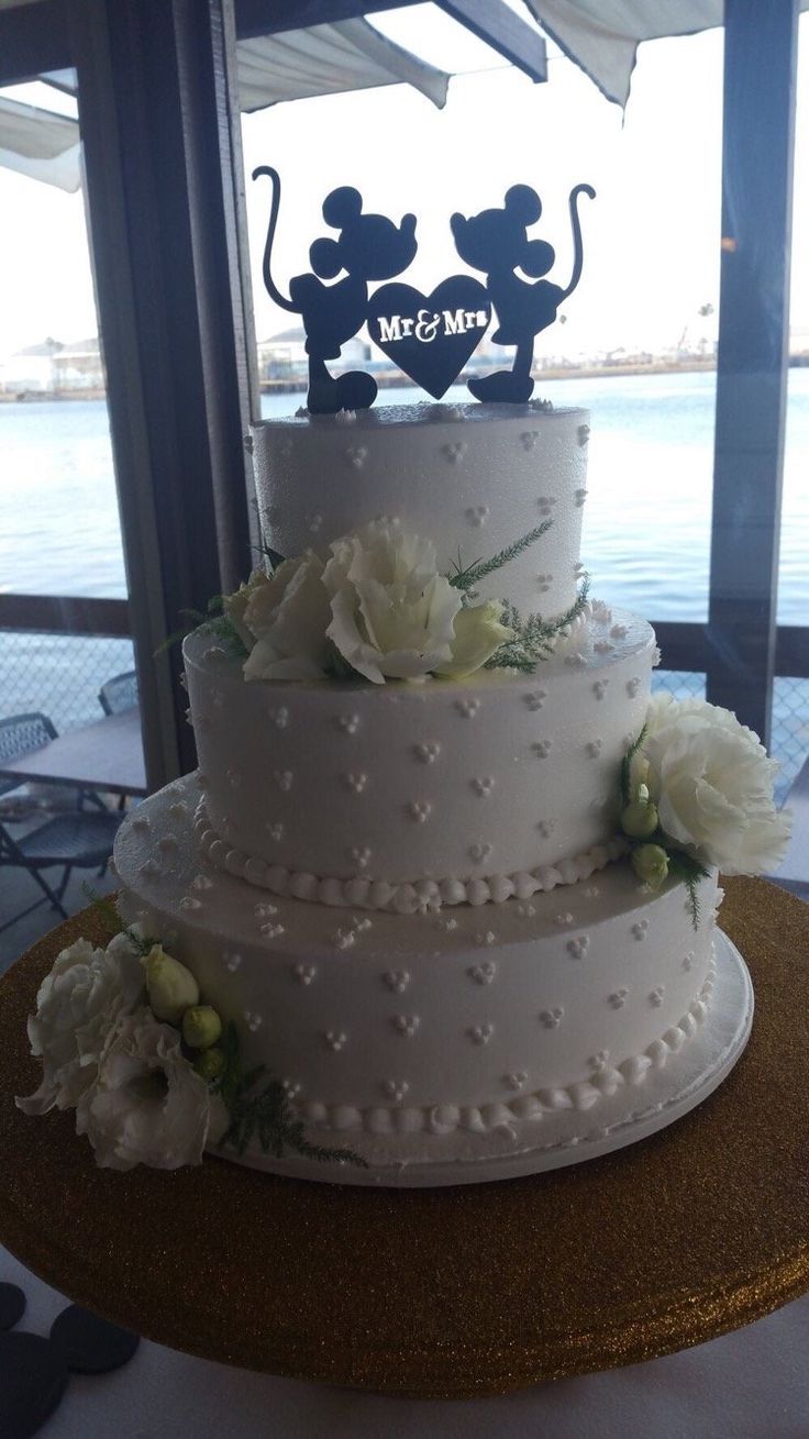 a three tiered wedding cake with white flowers and mickey mouse topper on it