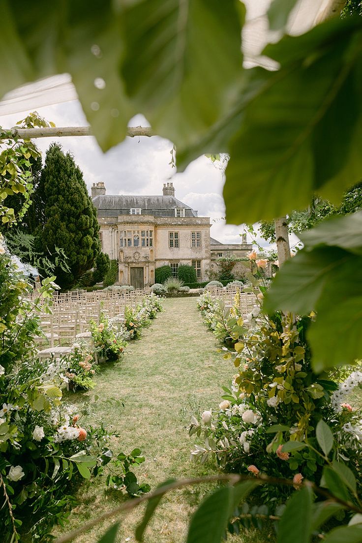 a large house surrounded by lush green trees and flowers in the foreground is a garden with lots of greenery