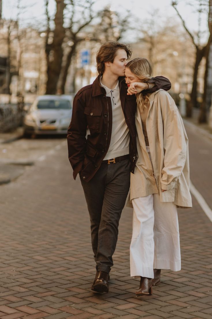 a young man and woman walking down the street in front of some trees, kissing