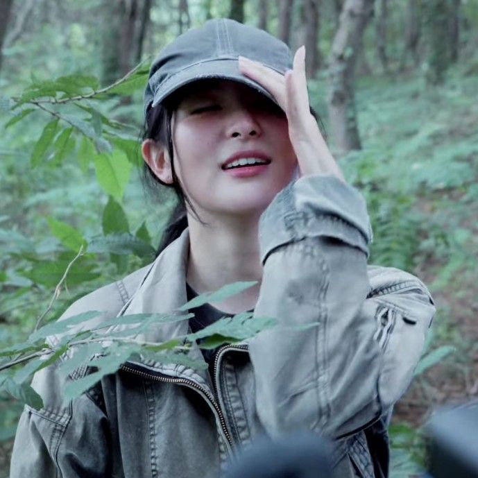 a woman wearing a hat in the woods with her hand up to her face and looking off into the distance