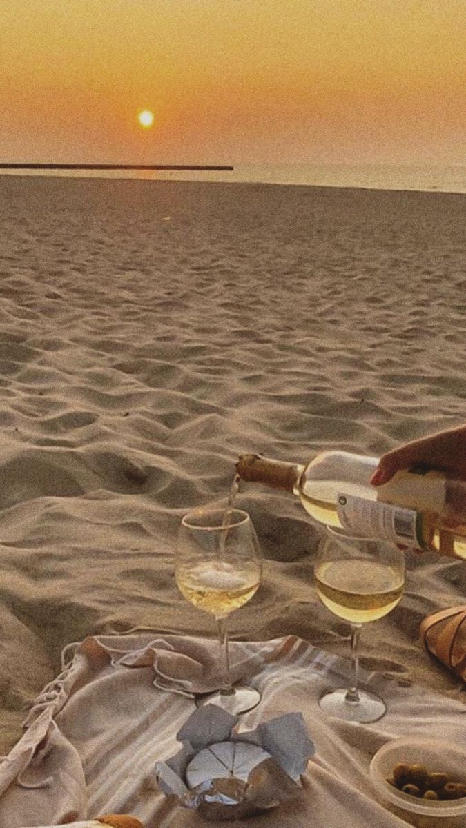 two glasses of wine are sitting on the beach with food and drinks in front of them