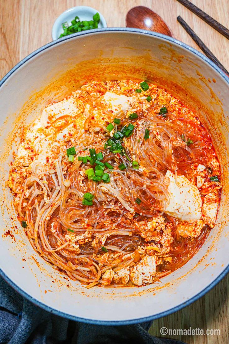 an overhead view of a bowl of noodles with meat and vegetables in tomato sauce on a wooden table